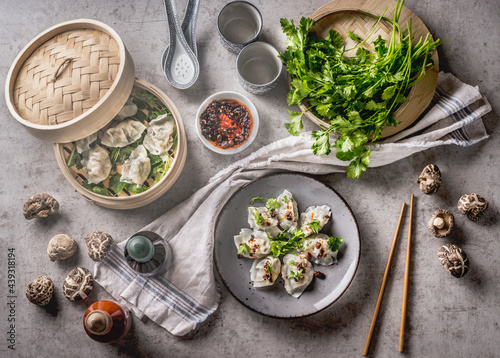 Asian food concept with homemade dumplings on plates and in steamer, fine shiitake, traditional sauces and crockery. White kitchen cloth on grey concrete background. Top view