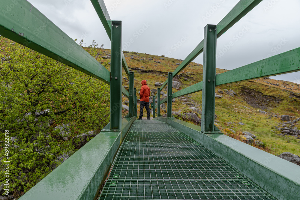 Man wearing orange jacket, standing at end of bridge in mountainous landscape in kerry ireland Man wearing orange jacket, standing at end of bridge in mountainous landscape in kerry ireland
