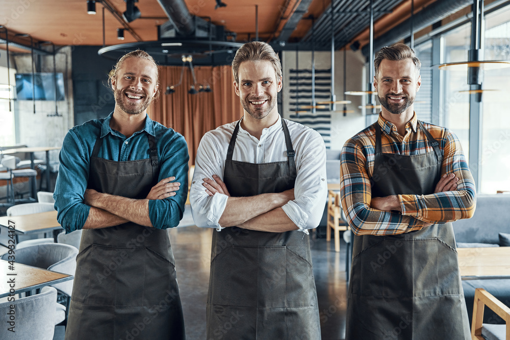 Three handsome male waiters in aprons keeping arms crossed and looking at camera