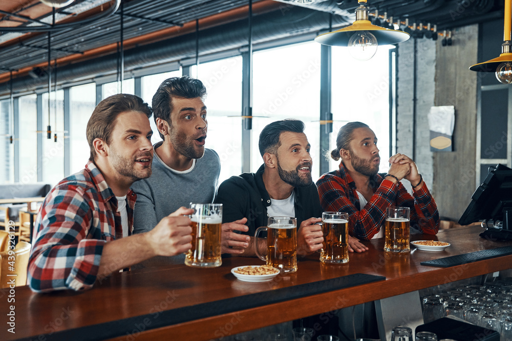 Cheering young men in casual clothing drinking beer and watching sport game while sitting in the pub Cheering young men in casual clothing drinking beer and watching sport game while sitting in the pub