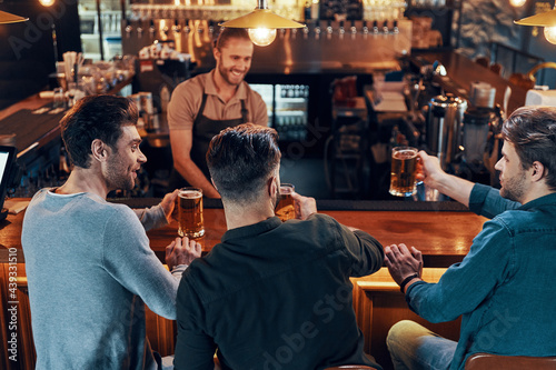 Smiling young men in casual clothing drinking beer while sitting in the pub together