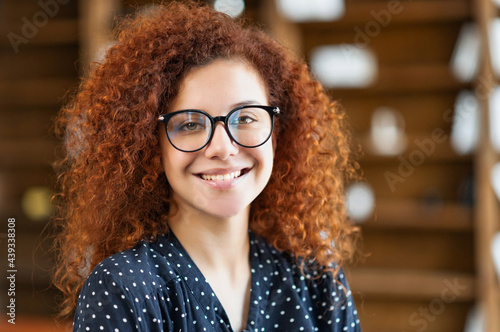 Headshot of young elegant female employee with curly ginger hairstyle, beautiful woman in stylish eyeglasses stands in office interior, smiles and looks at the camera, successful female entrepreneur