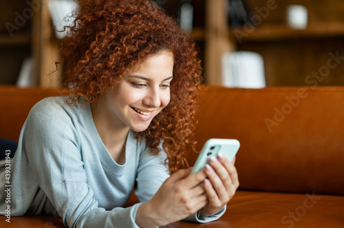 Happy young attractive curly redhead woman using mobile phone lying down on the cozy sofa, texting and messaging, sharing news in social media, cheerful girl enjoys chatting with friends online
