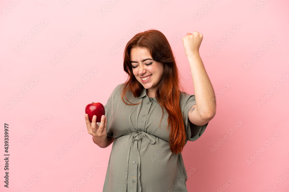 Young redhead caucasian woman isolated on pink background pregnant and holding an apple while celebrate a victory Young redhead caucasian woman isolated on pink background pregnant and holding an apple while celebrate a victory