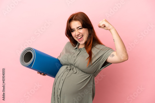 Young redhead caucasian woman isolated on pink background pregnant and doing strong gesture while going to yoga classes