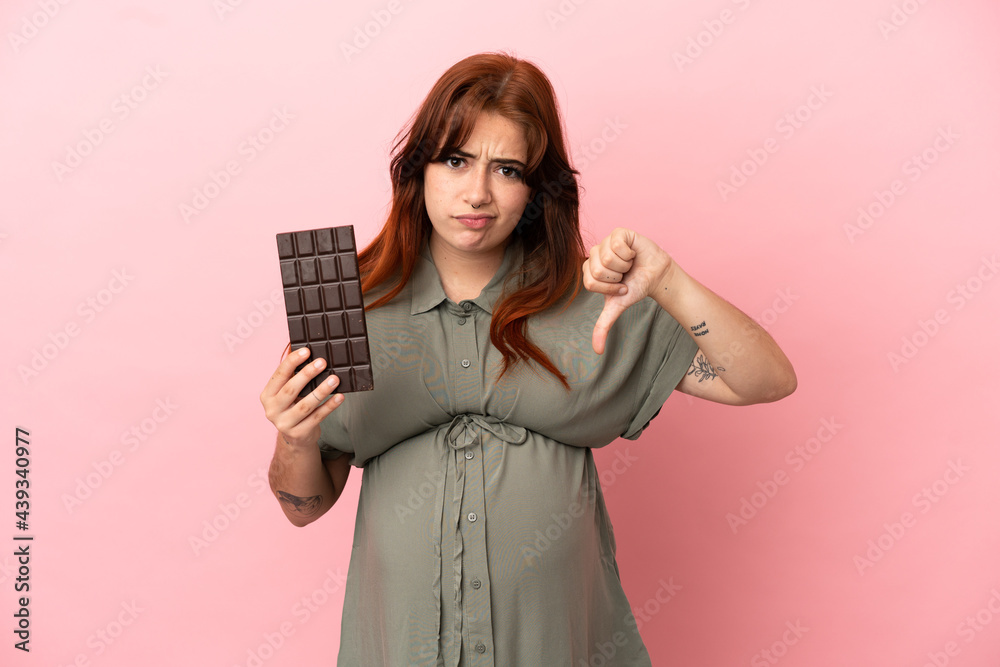 Young redhead caucasian woman isolated on pink background pregnant and holding chocolate doing bad signal Young redhead caucasian woman isolated on pink background pregnant and holding chocolate doing bad signal