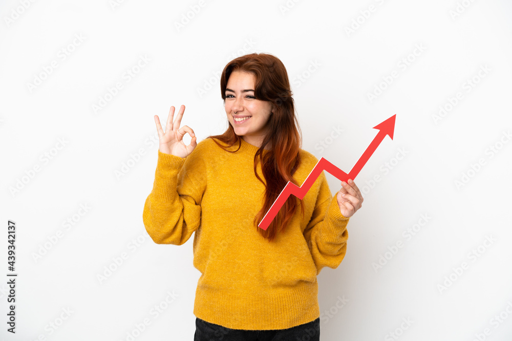 Young redhead woman isolated on white background holding a catching a rising arrow and doing OK sign Young redhead woman isolated on white background holding a catching a rising arrow and doing OK sign