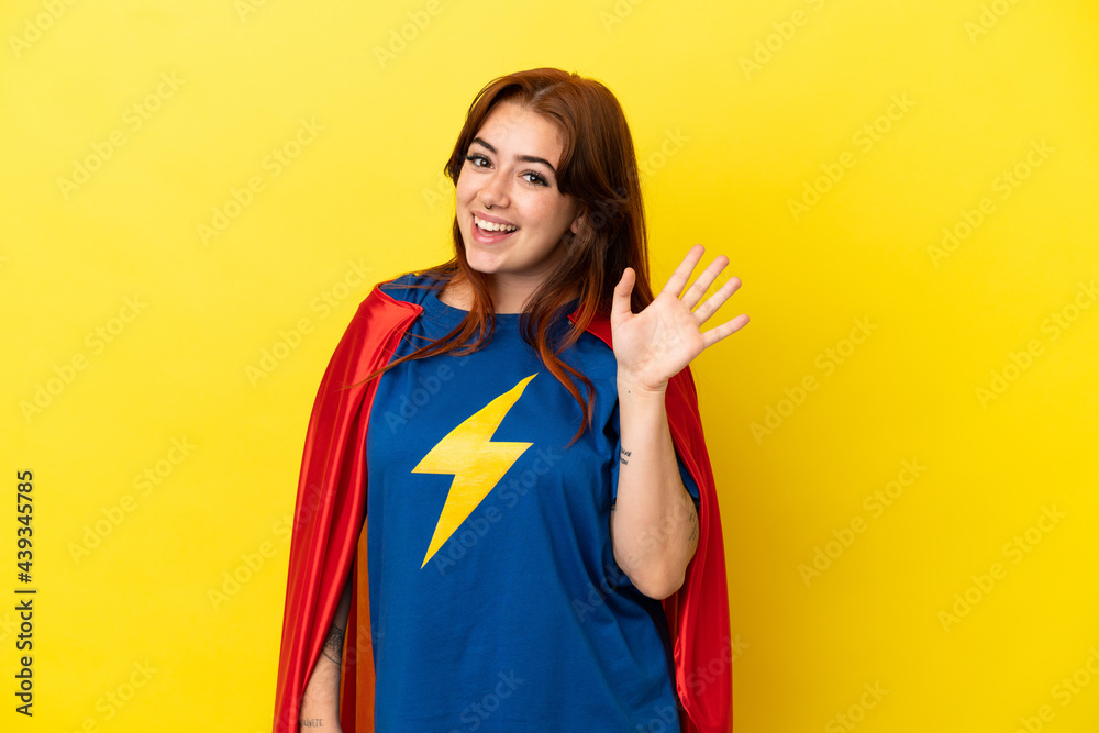 Super Hero redhead woman isolated on yellow background saluting with hand with happy expression