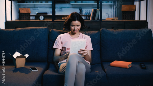 brunette young woman using digital tablet while sitting on sofa near book, smartphone and carton box with chinese food.