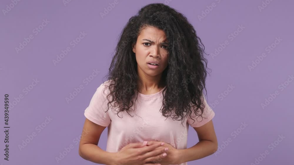 Sick ill sad tired young african woman 20s years old wears pink T-shirt put hands on abdomen suffer from stomach-ache griping bellyache feel bad seedy isolated on pastel violet color background studio Sick ill sad tired young african woman 20s years old wears pink T-shirt put hands on abdomen suffer from stomach-ache griping bellyache feel bad seedy isolated on pastel violet color background studio