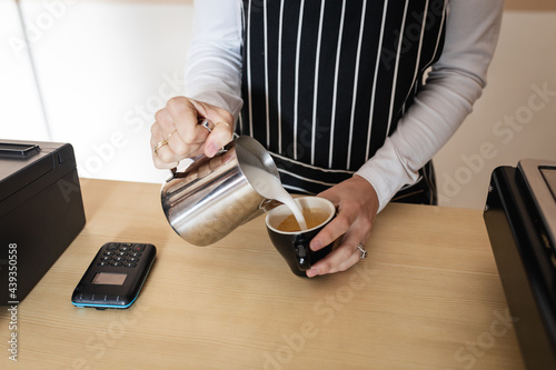 Midsection of caucasian female business owner wearing apron, pouring froth milk to coffee