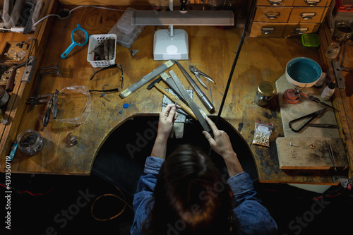 Caucasian female jeweller sitting at desk, holding jewelry tools, making jewelry in workshop photo