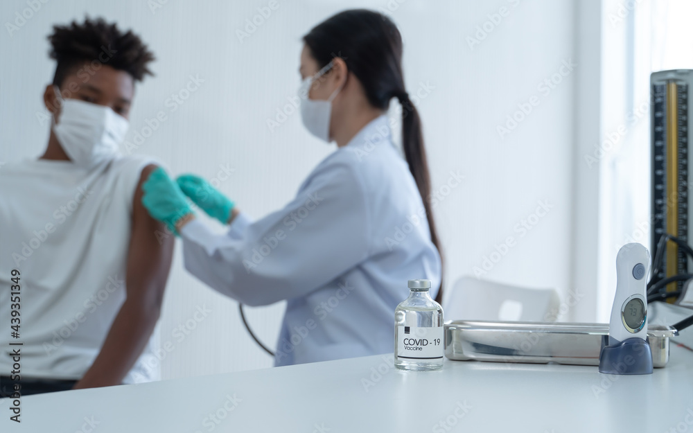 Fototapeta premium Selective focus on Covid-19 vaccine bottle on table. And next to it is thermometer and blood pressure monitor. Blur background is Asian woman doctor putting plaster on African boy arm after vaccinated
