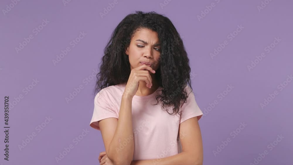 Puzzled young african american woman 20s years old wears pink T-shirt look aside put hand prop up chin iterates over solution options isolated on pastel violet purple color background studio portrait Puzzled young african american woman 20s years old wears pink T-shirt look aside put hand prop up chin iterates over solution options isolated on pastel violet purple color background studio portrait