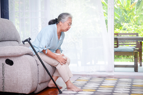 Asian elderly woman Sitting on the sofa in the house, there was a walking stick nearby. and had knee pain, unable to walk. concept of elderly health problems Prevention and health care in retirement.