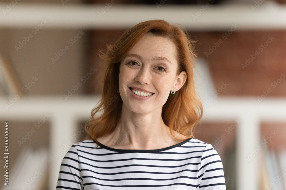 Head shot portrait of beautiful smiling young red-haired business lady posing in office. Sincere happy attractive millennial generation female employee worker entrepreneur expert looking at camera.