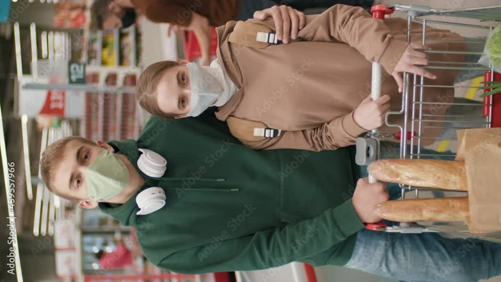 Vertical medium slow-motion portrait of happy young couple wearing masks smiling at camera after daily food shopping in supermarket standing next to cash desk