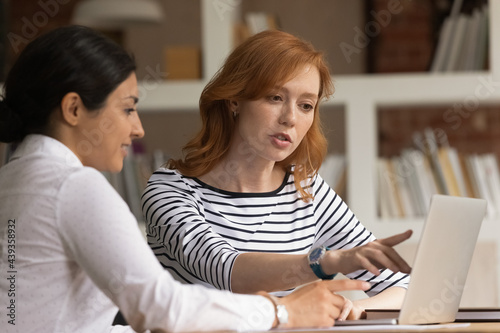 Concentrated young red-haired business lady discussing online project or analyzing sales data statistics with indian colleague in office. Focused young mixed race employees working on computer.