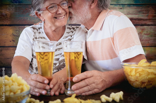 Bearded senior man kisses his wife sitting at pub at a wooden table toasting with two glasses of beer and potato chips. Happy relaxed retired couple