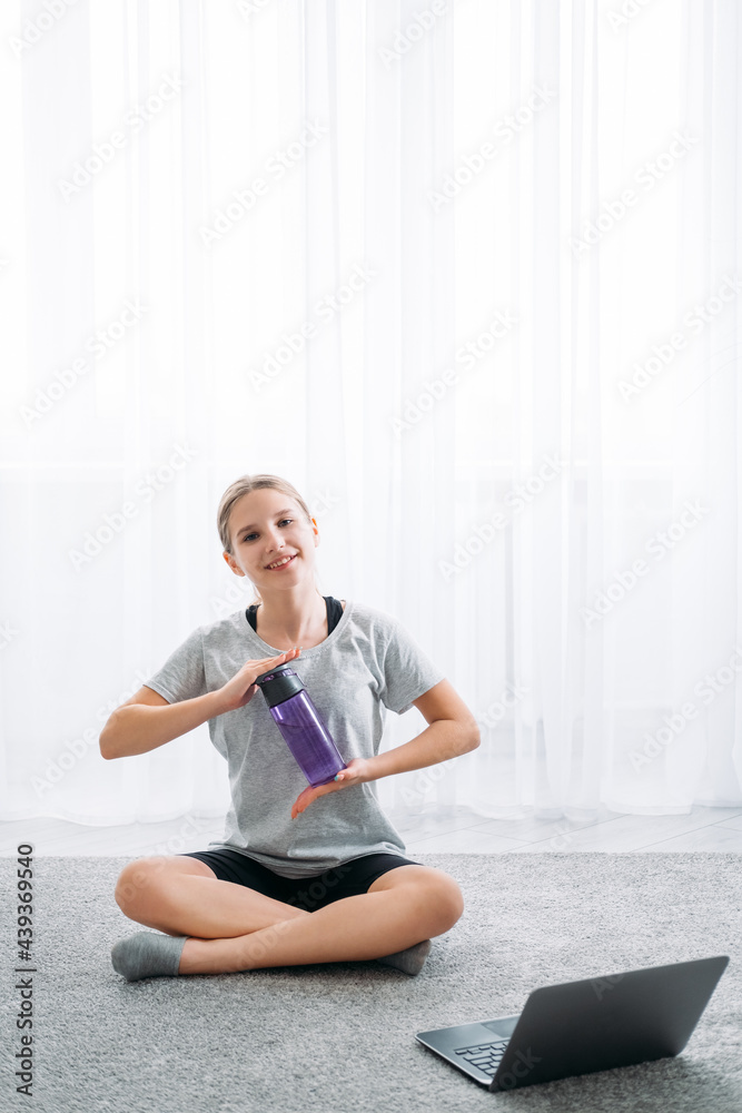 Home sport. Fitness refreshing. Online training. Happy girl sitting cross-legged on floor with laptop holding water bottle on light white window copy space background. Home sport. Fitness refreshing. Online training. Happy girl sitting cross-legged on floor with laptop holding water bottle on light white window copy space background.