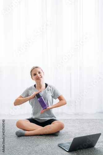 Home sport. Fitness refreshing. Online training. Happy girl sitting cross-legged on floor with laptop holding water bottle on light white window copy space background.