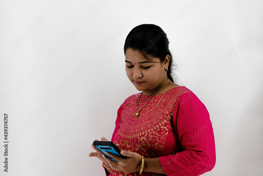 Smiling happy young indian ethnicity lady standing on right, aside blank empty free copy space for advertising text, looking at mobile phone screen, receiving good offer isolated on white background.. Smiling happy young indian ethnicity lady standing on right, aside blank empty free copy space for advertising text, looking at mobile phone screen, receiving good offer isolated on white background..
