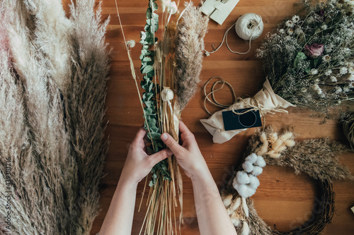 Top View of Female Hands Making Everlasting Bouquet of Dried Flowers at Wooden Table with Bunch of Flowers and Decorations at her Flower Shop Studio photo