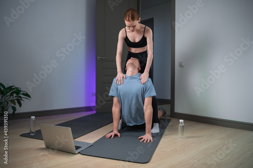 The wife conducts yoga training at home with her husband, she helps him to perform the exercises correctly, the husband performs the upward-facing dog asana