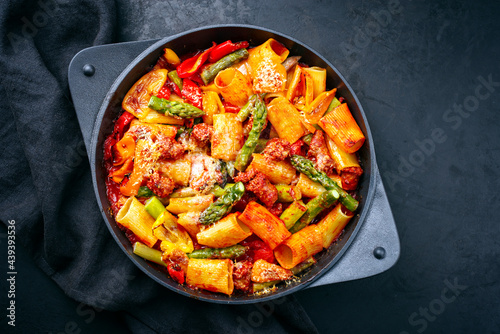 Traditional roasted Italian pasta mezzi paccheri rigati con salsiccia sausage with green asparagus tips in tomato sauce served as top view in cast iron skillet pan in matte flat cinematic modern look