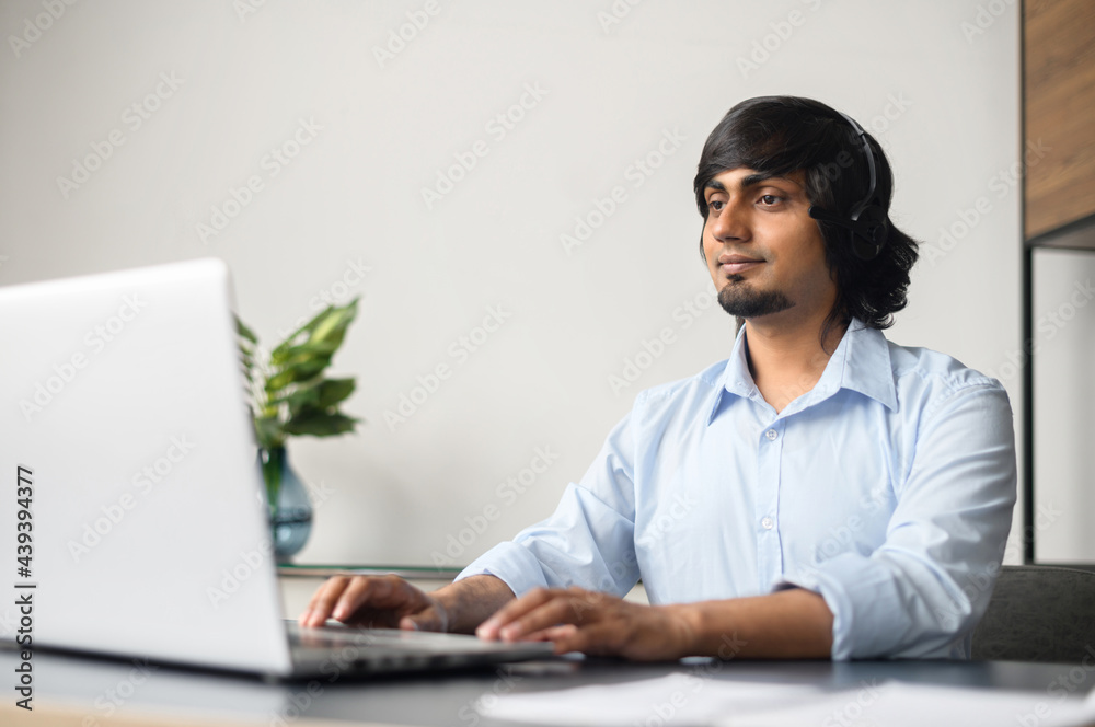 Smiling indian businessman wearing headset is using laptop computer for online communication with customers or colleagues sitting in the modern office Smiling indian businessman wearing headset is using laptop computer for online communication with customers or colleagues sitting in the modern office