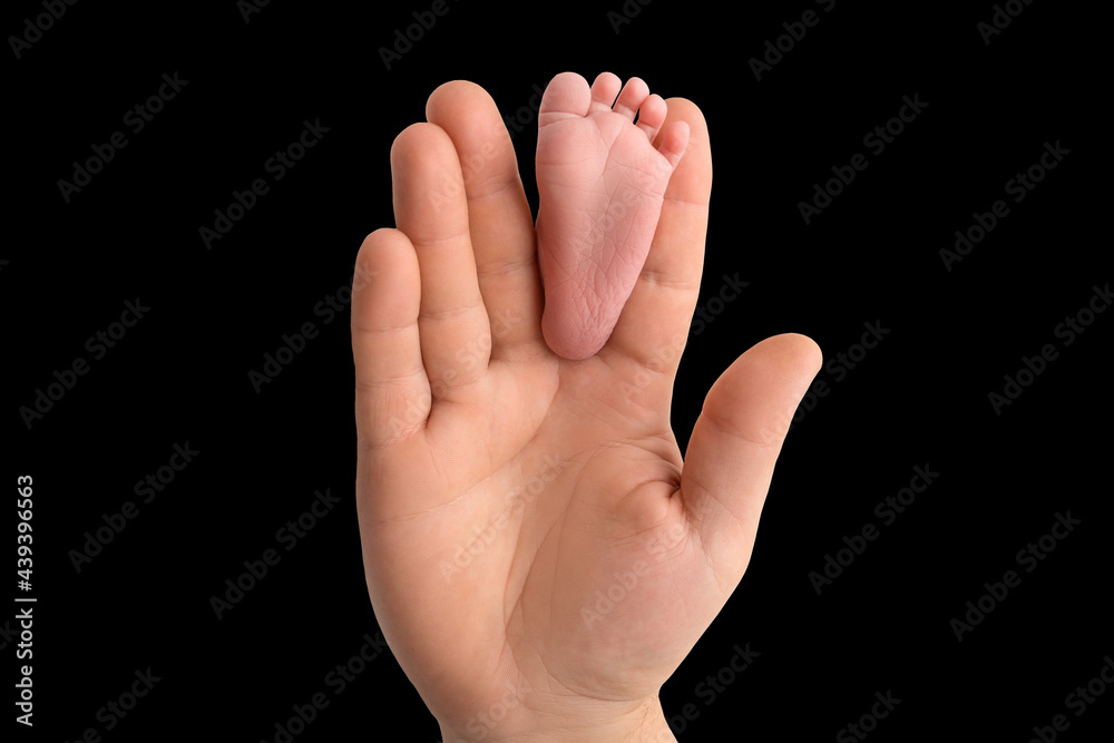 Baby feet in father's hand. The tiny foot of a newborn baby between the fingers of a parent's hand. Open palm and small toes. Black studio background. Baby feet in father's hand. The tiny foot of a newborn baby between the fingers of a parent's hand. Open palm and small toes. Black studio background.