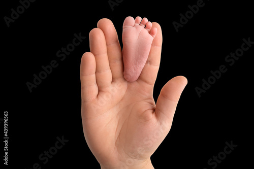 Baby feet in father's hand. The tiny foot of a newborn baby between the fingers of a parent's hand. Open palm and small toes. Black studio background. © Vad-Len