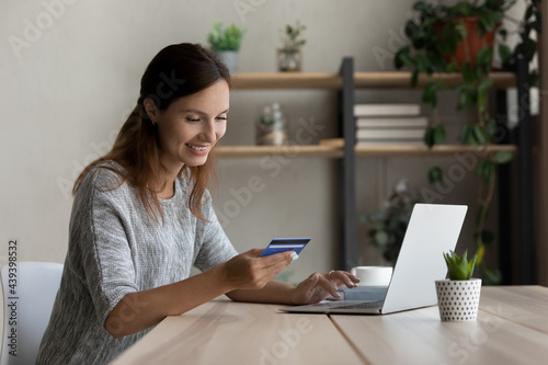 Happy woman holding plastic credit card, using laptop, paying online, smiling satisfied young female customer making secure internet payment, entering information, typing, browsing banking service