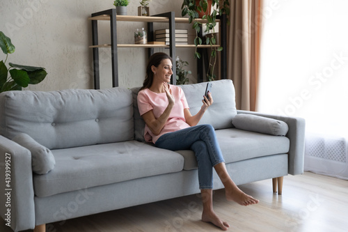 Smiling Arabian woman holding smartphone, waving hand, making video call to friends or relatives, sitting on couch at home, chatting in social network online, blogger influencer greeting viewers