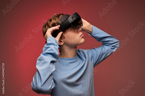 shocked young guy in virtual reality helmet, looking into VR glasses screen. Young caucasian boy is surprised by reality VR helmet. Isolated in studio on red background, portrait. modern technologies