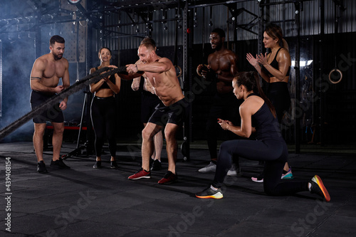 Strong shirtless man engaged in cross fit battling ropes at gym workout exercise, while supportive group of friends stand next to him, in sportive outfit
