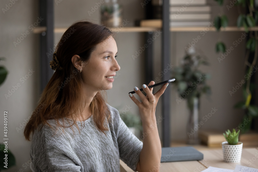 Side view smiling woman recording audio message on smartphone, chatting in social network, positive attractive young woman using phone, making call, talking on loudspeaker, activating voice assistant Side view smiling woman recording audio message on smartphone, chatting in social network, positive attractive young woman using phone, making call, talking on loudspeaker, activating voice assistant
