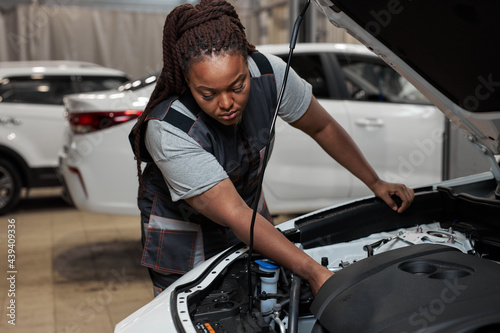 Black woman mechanic working under the hood at repair garage. Portrait of confident focused mechanic woman working ona car in an auto repair shop. Female mechanic working on car. Side view