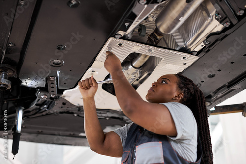 Black Female Auto Mechanic Working Underneath Car In Garage. Portrait of hardworking young female mechanic in overalls, inspecting lifted car. Mechanic woman working in car repair shop. Side View