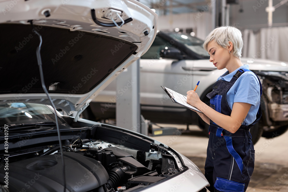 Woman auto Mechanic writing on clipboard at repair garage, wearing uniform overalls. Young mechanic engineer female taking notes on clipboard for examining a vehicle, side view Woman auto Mechanic writing on clipboard at repair garage, wearing uniform overalls. Young mechanic engineer female taking notes on clipboard for examining a vehicle, side view