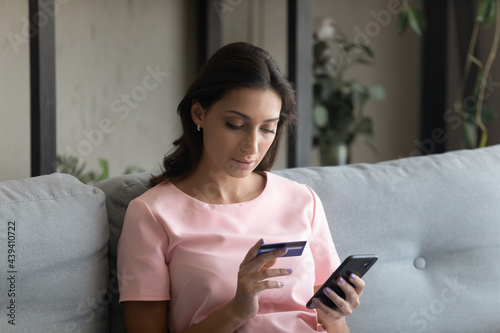 Close up focused Arabian woman holding credit card and smartphone entering information, sitting on couch at home, young female customer paying online, shopping in internet store, checking balance