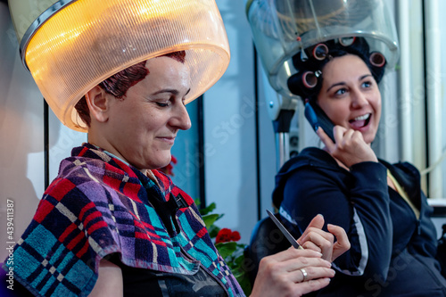 Two female friends at the hairdresser in a beauty salon sitting under a vintage hood hair dryer, talking, gossiping, and laughing while doing their nails. Retro style and fashion. Selective focus. photo