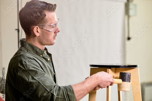 Handsome joiner working with hammer in furniture workshop, making wooden chair, side view portrait of focused guy in green shirt, indoors. © Roman