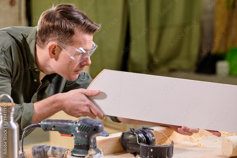 Side view portrait of caucasian joiner in protective goggles checking the surface of wooden board material in workshop, in green shirt, looking confident.