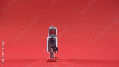 A tin toy robot, walking over a red backdrop, losing power until it reaches its discharge point and it stops. Profile shot.
