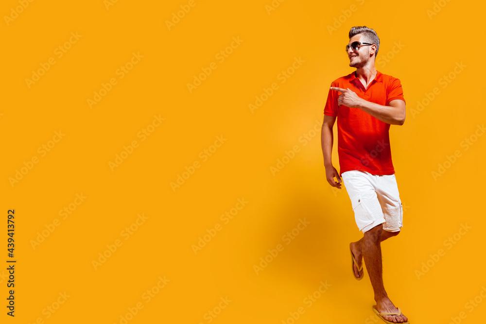 Full length portrait of a handsome man in shorts and red shirt walking and looking sideway, pointing finger isolated on yellow background in studio. Full length portrait of a handsome man in shorts and red shirt walking and looking sideway, pointing finger isolated on yellow background in studio.