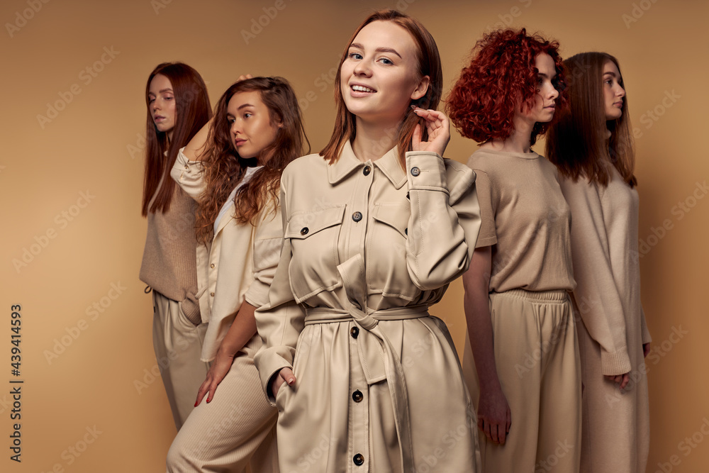 Redhead Smiling female in trench coat posing at camera standing among female models in the background, portrait. Isolated beige background. fashion, style, trend concept
