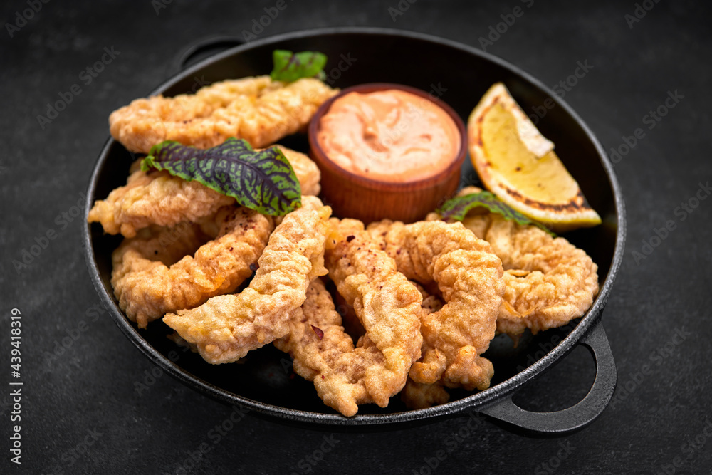 Golden rings of squid fries in a round frying pan with sauce, herbs and lemon on a dark background without decorations Golden rings of squid fries in a round frying pan with sauce, herbs and lemon on a dark background without decorations