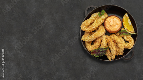 Golden rings of squid fries in a round frying pan with sauce, herbs and lemon on a dark background without decorations