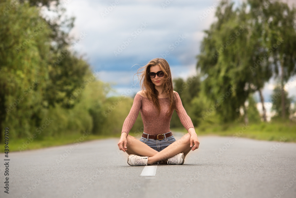 Sexy young woman in sunglasses sits on the asphalt road.